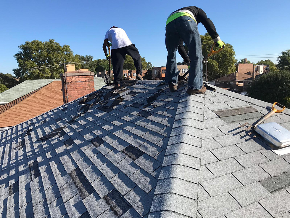 Two men work on a gray shingled roof with houses in distance
